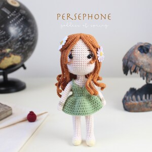 Puede incluir: Muñeca de ganchillo hecha a mano llamada Perséfone, la diosa de la primavera, con cabello rojo y un vestido verde. La muñeca tiene acentos de margaritas blancas en su cabello. El fondo incluye un globo terráqueo y un cráneo de dinosaurio.