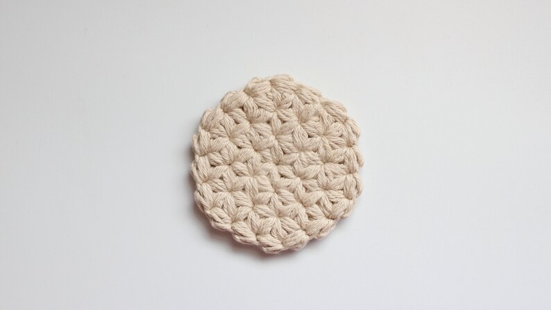 May include: A circular coaster crafted from thick, off-white cotton yarn. The piece features a repeating star-like pattern created through dense crochet stitching, resulting in a textured, raised surface with a scalloped, uneven outer edge.