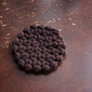 May include: A circular dark brown coaster featuring a dense, textured crochet pattern made of interconnected yarn loops. The coaster has a thick, padded appearance with a repeating star-like stitch design across its entire surface.