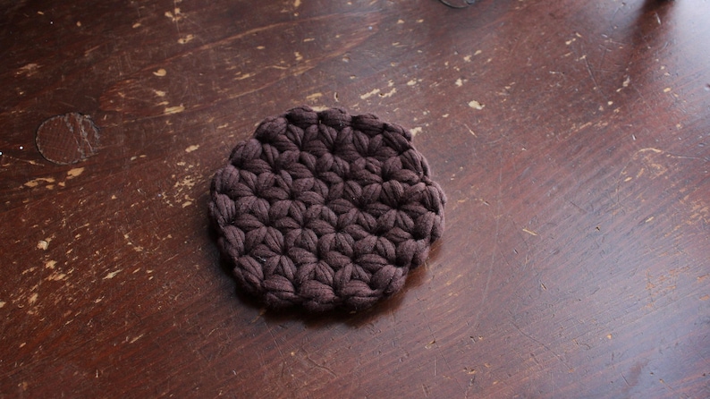 May include: A circular dark brown coaster featuring a dense, textured crochet pattern made of interconnected yarn loops. The coaster has a thick, padded appearance with a repeating star-like stitch design across its entire surface.