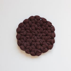 May include: A circular coaster crafted from dark brown yarn, featuring a dense, repeating puff stitch pattern that creates a textured, floral-like appearance across the entire surface of the round, thick textile object.