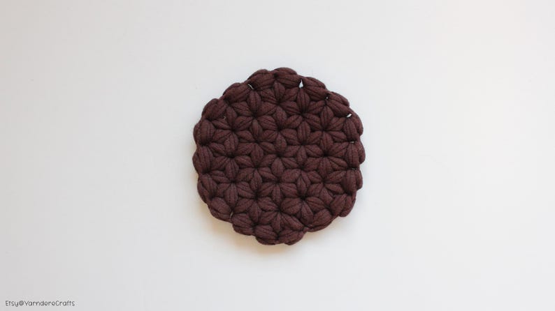 May include: A circular coaster crafted from dark brown yarn, featuring a dense, repeating puff stitch pattern that creates a textured, floral-like appearance across the entire surface of the round, thick textile object.