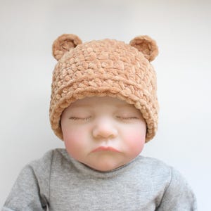 May include: A tan baby hat with bear ears, made from a soft, textured material. The baby is wearing a grey long-sleeved top. The baby's eyes are closed.