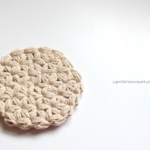 May include: A round, beige crocheted coaster featuring a textured star-like pattern made from thick cotton yarn. The text "Light/DK/Worsted/8-ply cotton yarn" is printed to the right of the coaster, indicating the material used for the item.