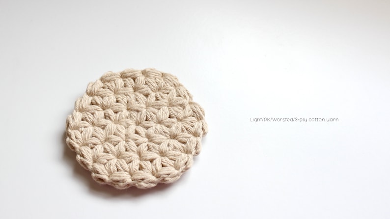 May include: A round, beige crocheted coaster featuring a textured star-like pattern made from thick cotton yarn. The text "Light/DK/Worsted/8-ply cotton yarn" is printed to the right of the coaster, indicating the material used for the item.