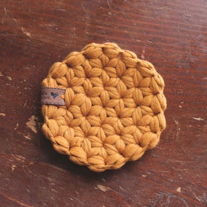 May include: A round, mustard-yellow crocheted coaster featuring a textured, star-like pattern throughout. A small, rectangular brown faux-leather tag is attached to the side, displaying a small heart icon and partially visible text.