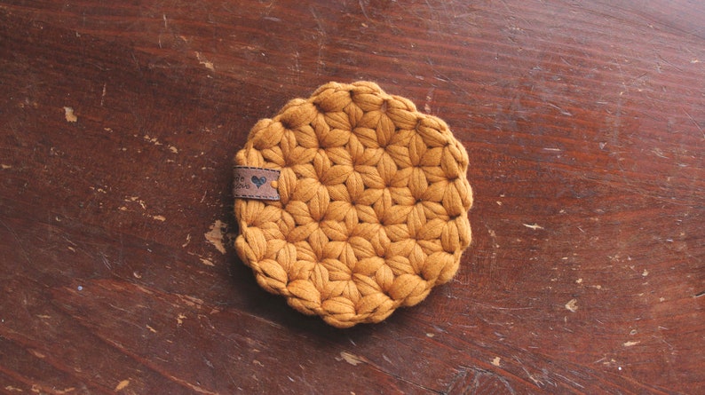 May include: A round, mustard-yellow crocheted coaster featuring a textured, star-like pattern throughout. A small, rectangular brown faux-leather tag is attached to the side, displaying a small heart icon and partially visible text.