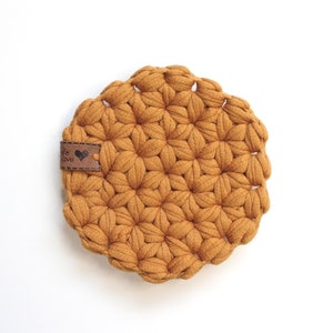 Op de afbeelding: Een ronde, mosterdgele gehaakte onderzetter met een bruin leren label waarop "Met liefde gemaakt" staat.