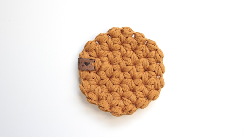 May include: A round, mustard-yellow crocheted coaster featuring a textured, repeating star-like pattern. A small, rectangular brown leather tag is attached to the left side, displaying a heart icon and the partially visible text "made with love."