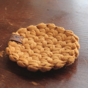 May include: A round, mustard-yellow crochet coaster featuring a textured, puff-stitch pattern. A small, rectangular brown leather tag is attached to the edge, displaying a tiny blue heart icon and some indistinct, small black text.