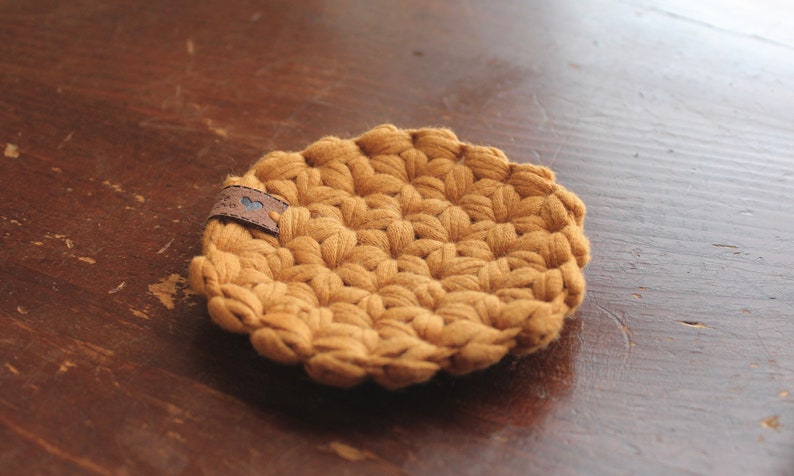 May include: A round, mustard-yellow crochet coaster featuring a textured, puff-stitch pattern. A small, rectangular brown leather tag is attached to the edge, displaying a tiny blue heart icon and some indistinct, small black text.