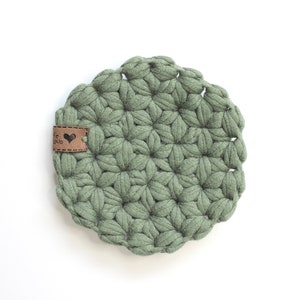 May include: A round, sage green crocheted coaster featuring a textured, star-like pattern made from thick yarn. A small, rectangular brown leather tag is attached to the left side, displaying a black heart icon and some illegible text.