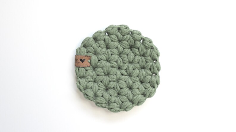 May include: A round, sage green crocheted coaster featuring a textured, star-like pattern made from thick yarn. A small, rectangular brown leather tag is attached to the left side, displaying a black heart icon and some illegible text.