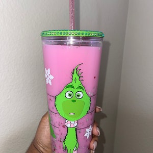 K&ouml;nnte beinhalten: Ein wiederverwendbarer Becher in Pink und Gr&uuml;n mit einem gr&uuml;nen Grinch-Charakterdesign und einem rosa glitzernden Deckel. Der Becher hat einen durchsichtigen Plastikstrohhalm.