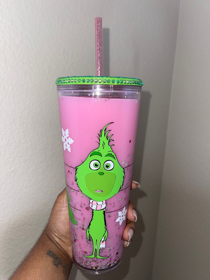 K&ouml;nnte beinhalten: Ein wiederverwendbarer Becher in Pink und Gr&uuml;n mit einem gr&uuml;nen Grinch-Charakterdesign und einem rosa glitzernden Deckel. Der Becher hat einen durchsichtigen Plastikstrohhalm.