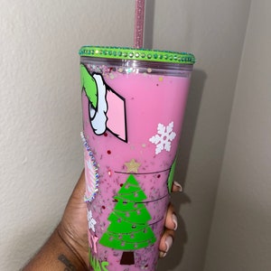 K&ouml;nnte beinhalten: Ein wiederverwendbarer Becher in Pink und Gr&uuml;n mit einem Weihnachtsbaum- und Grinch-Design. Der Becher hat einen durchsichtigen Deckel mit einem gr&uuml;nen Glitzerrand und einem rosa Strohhalm.