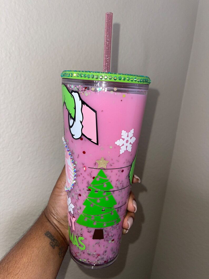 K&ouml;nnte beinhalten: Ein wiederverwendbarer Becher in Pink und Gr&uuml;n mit einem Weihnachtsbaum- und Grinch-Design. Der Becher hat einen durchsichtigen Deckel mit einem gr&uuml;nen Glitzerrand und einem rosa Strohhalm.