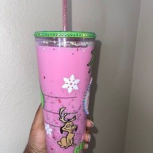 K&ouml;nnte beinhalten: Ein wiederverwendbarer Becher in Pink und Gr&uuml;n mit einem gr&uuml;nen Deckel und einem rosa Strohhalm. Der Becher hat ein Grinch-Charakterdesign mit einem Schneeflocke und dem Wort "Grinch" darauf. Der Becher ist mit einer rosa Fl&uuml;ssigkeit gef&uuml;llt und enth&auml;lt Glitzer.