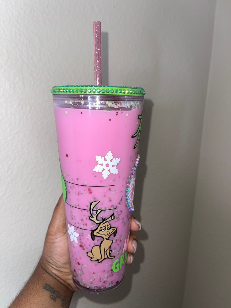 K&ouml;nnte beinhalten: Ein wiederverwendbarer Becher in Pink und Gr&uuml;n mit einem gr&uuml;nen Deckel und einem rosa Strohhalm. Der Becher hat ein Grinch-Charakterdesign mit einem Schneeflocke und dem Wort "Grinch" darauf. Der Becher ist mit einer rosa Fl&uuml;ssigkeit gef&uuml;llt und enth&auml;lt Glitzer.