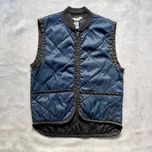 May include: A navy blue quilted vest with a black zipper and black trim. The vest has a diamond quilted pattern and a ribbed collar.
