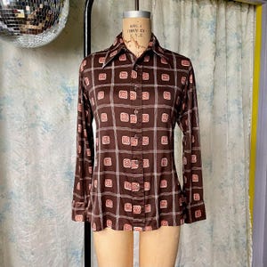 May include: A brown long-sleeved shirt with a white and pink geometric pattern. The shirt has a collar and button closure.