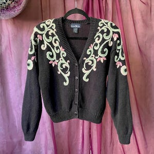 May include: A black cardigan sweater with a V-neck and button closure. The sweater features an embroidered floral design in light green and pink on the shoulders and upper chest. The sweater is hanging on a black hanger.