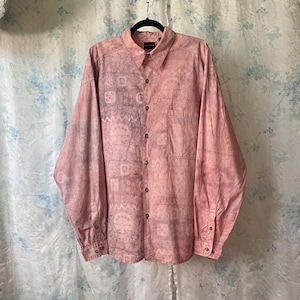 May include: A long-sleeved button-down shirt in a faded pink hue, featuring a subtle, patterned design. The shirt has a single chest pocket and is hanging on a black hanger. The fabric appears soft and lightweight.