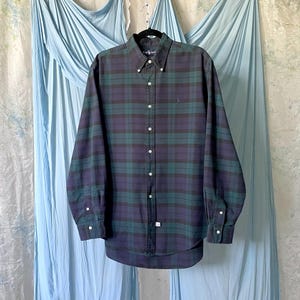 May include: A long-sleeve button-down shirt featuring a dark green and navy blue plaid pattern. The shirt has a button-down collar, a front button placket, and buttoned cuffs. A small embroidered logo is visible on the left chest area.
