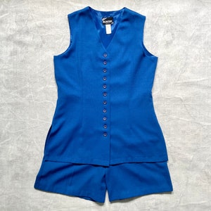 Op de afbeelding: Een koninklijkblauwe, mouwloze vest met een knoopsluiting aan de voorkant en bijpassende shorts. De vest heeft een V-hals en de shorts hebben een rechte pijp.