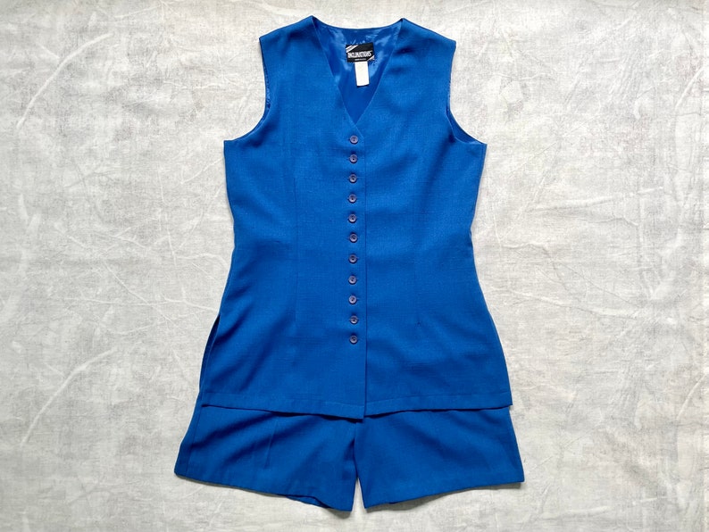 Op de afbeelding: Een koninklijkblauwe, mouwloze vest met een knoopsluiting aan de voorkant en bijpassende shorts. De vest heeft een V-hals en de shorts hebben een rechte pijp.