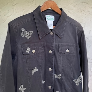 May include: A dark gray denim jacket adorned with sparkling rhinestone butterfly designs. The jacket features a classic collar, button-down front, and silver buttons. A stylish outerwear piece.