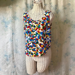 May include: A sleeveless top with a vibrant, abstract pattern in red, blue, yellow, green, and white. The top is displayed on a wicker mannequin. The design features geometric shapes and floral-like motifs, creating a colorful and eye-catching garment.