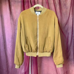 May include: A tan bomber jacket with a full-length zipper, two front pockets, and ribbed cuffs and hem. The jacket is hanging on a white hanger against a pink and purple backdrop.