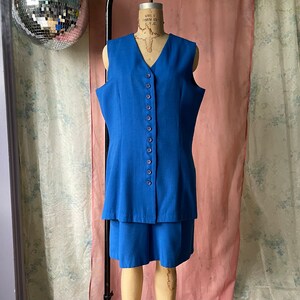Op de afbeelding: Een blauwe, mouwloze jurk met een knoopsluiting en een rok met een gelaagde look.