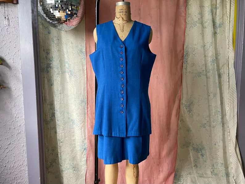 Op de afbeelding: Een blauwe, mouwloze jurk met een knoopsluiting en een rok met een gelaagde look.