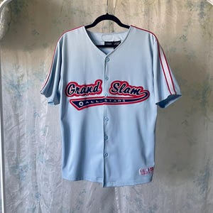 May include: Light blue baseball jersey with the words "Grand Slam All-Stars" in red and white lettering across the chest. The jersey has a V-neck, button-up front, and red and white stripes on the sleeves.