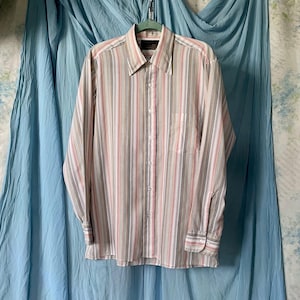 May include: A button-down shirt with vertical stripes in shades of beige, gray, pink, and white. The long-sleeved shirt has a collared neckline and a chest pocket. The shirt is hanging on a green hanger against a blue backdrop.