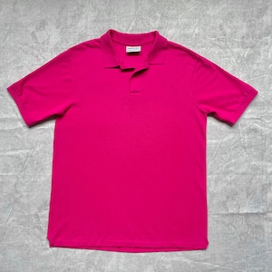 May include: A solid magenta polo shirt with short sleeves and a collared neckline. The shirt has a two-button placket and is made of a smooth, likely cotton, fabric. The shirt is laid flat on a textured, light gray surface.