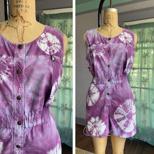 Op de afbeelding: Paarse en witte tie-dye jumpsuit zonder mouwen met een knoopsluiting aan de voorkant en een trekkoord in de taille.