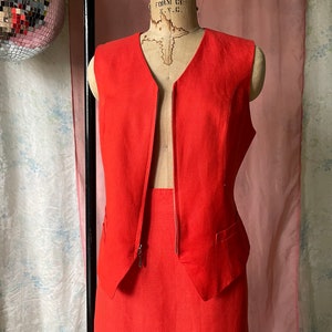 May include: A red sleeveless linen waistcoat with a deep V-neck and a zip fastening. The waistcoat is paired with a matching red skirt.
