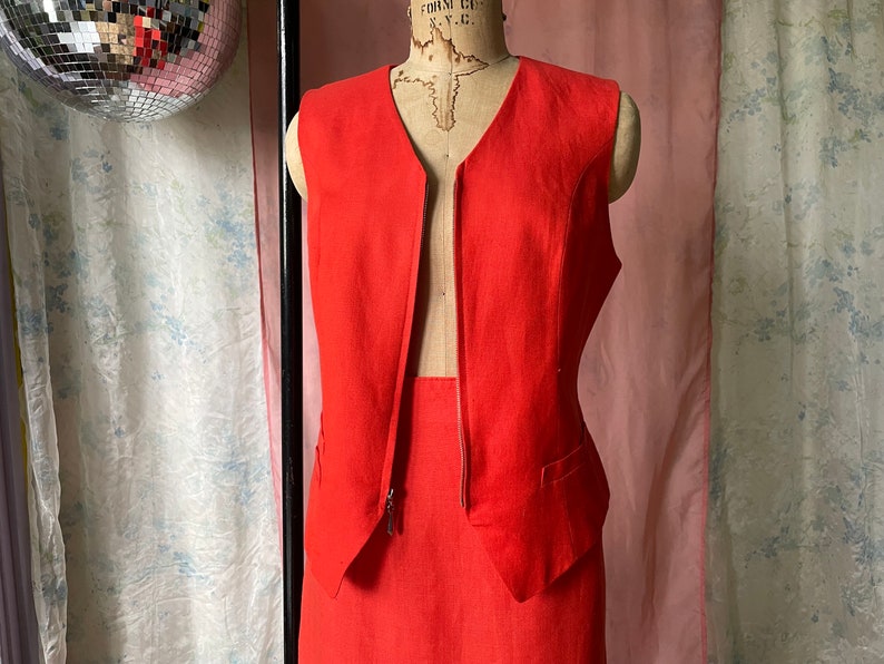 May include: A red sleeveless linen waistcoat with a deep V-neck and a zip fastening. The waistcoat is paired with a matching red skirt.