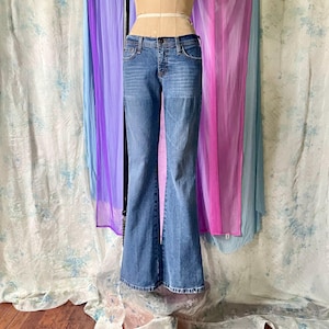 May include: Light blue denim jeans with a flared leg design. The jeans have a classic five-pocket style and a button and zipper closure. The jeans are displayed on a mannequin. The background includes sheer fabric in shades of purple, pink, and blue.