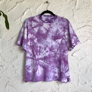 Op de afbeelding: Paarse tie-dye T-shirt met een zakje aan de linkerkant. Het shirt heeft een witte achtergrond met paarse swirls.