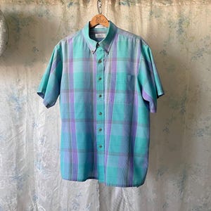 May include: A short-sleeved button-down shirt in a plaid pattern of teal, lavender, and gray. The shirt has a button-down collar and a single chest pocket. The shirt is hanging on a wooden hanger.