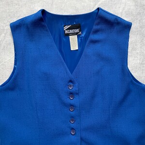 Op de afbeelding: Een koninklijk blauwe, mouwloze vest met een V-hals en vijf knopen aan de voorkant. De vest heeft een geweven textuur en een label met de tekst "Inclinations".