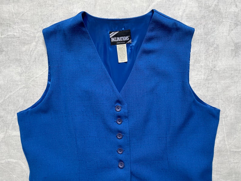 Op de afbeelding: Een koninklijk blauwe, mouwloze vest met een V-hals en vijf knopen aan de voorkant. De vest heeft een geweven textuur en een label met de tekst "Inclinations".
