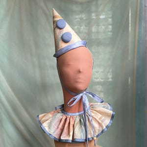 May include: A conical party hat and a matching ruffled collar. The hat is beige with a subtle leaf pattern, two blue circular accents, and a blue ribbon trim. The collar features the same beige leaf print with blue ribbon edges and ties.