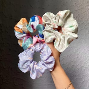 May include: Three fabric hair scrunchies with a smooth, shiny finish. One scrunchie features a multi-colored abstract pattern of orange, blue, and pink. Another is a solid light lavender, and the third is a cream color with subtle pastel accents.