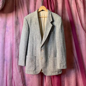 May include: A light gray tweed blazer with two front pockets and two buttons. The blazer is hanging on a wooden hanger against a pink background. The blazer has a notched lapel and a light beige lining.