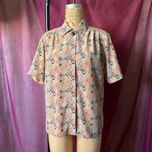 May include: Short-sleeved button-down shirt with a geometric pattern in peach, black, and purple. The shirt is displayed on a mannequin with a neutral-toned neck and shoulders. The fabric has a subtle sheen.